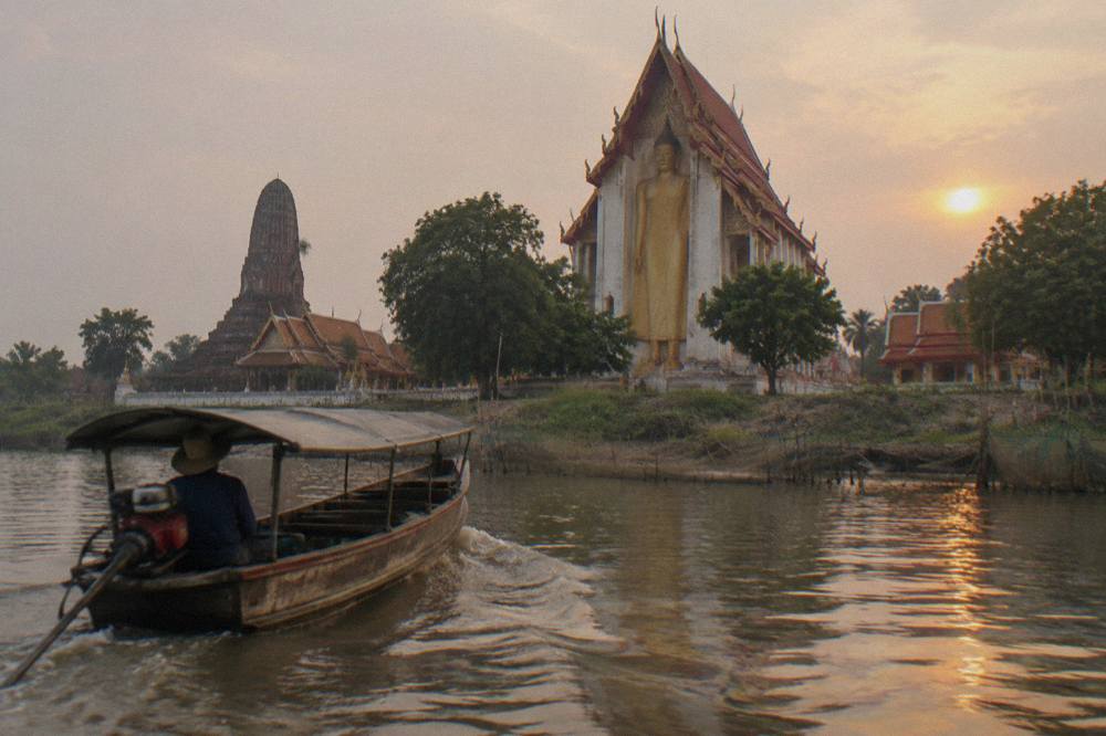 A wooden river boat passing temple ruins on the riverbank at golden hour