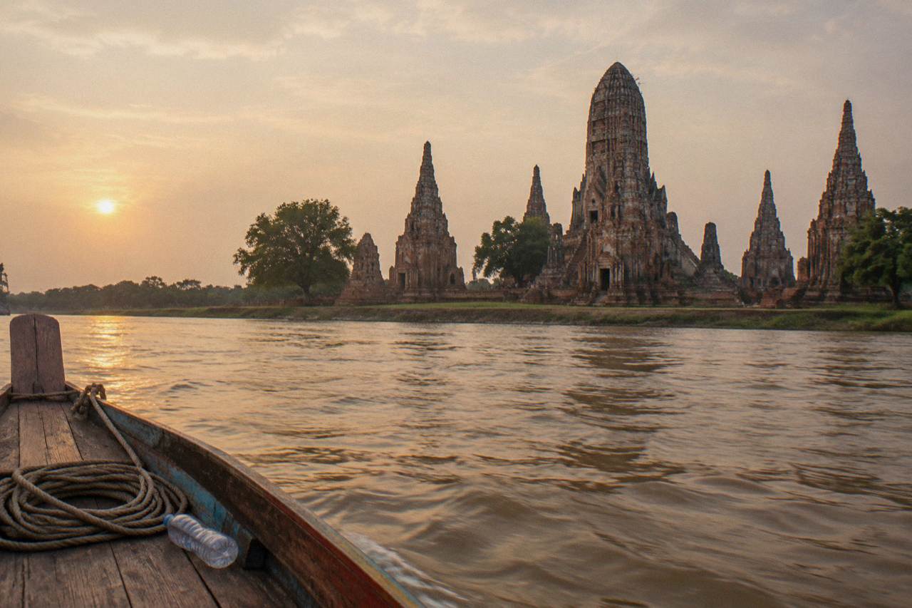 A longtail boat drifting past temple ruins beside a wide river at golden hour