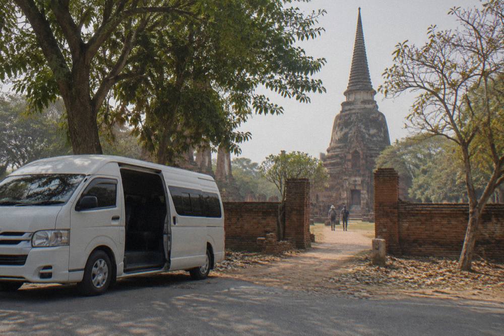 Air-conditioned tour van parked on a tree-lined road beside old brick ruins