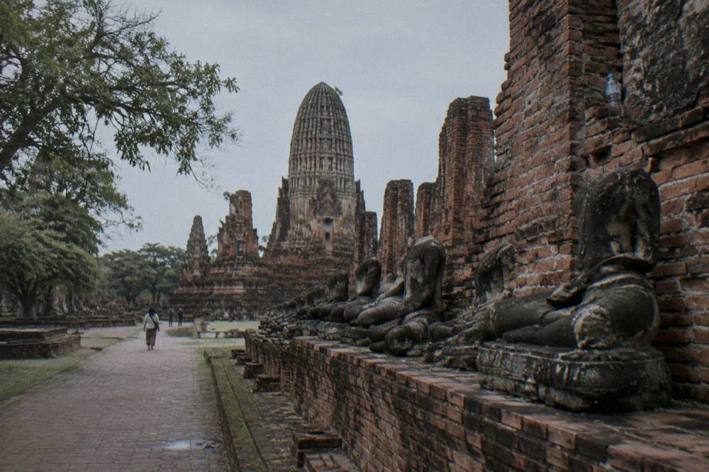 A cyclist on a quiet paved road approaching weathered brick temple ruins scattered with headless Buddha statues