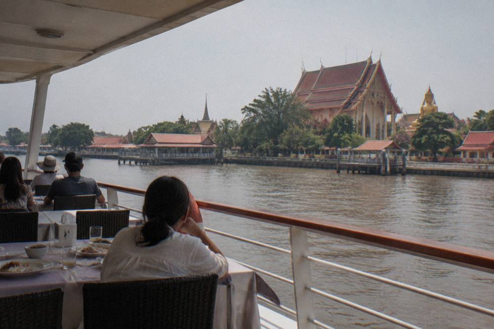 A large double-decker river cruise boat on the Chao Phraya River with passengers dining on the upper deck