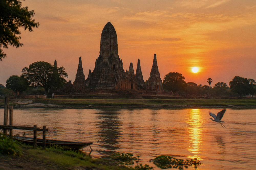 Sunset over Wat Chaiwatthanaram with the central prang silhouetted against an orange sky, seen from the river