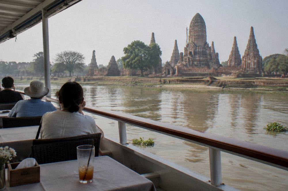 Passengers seated at tables on the upper deck of a river cruise boat, watching the Chao Phraya riverbank slide by