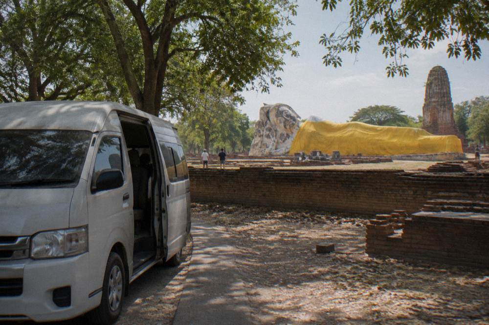 A small tour van parked on a shaded road beside ancient brick temple ruins
