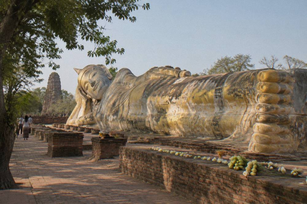 A long reclining Buddha statue resting in open-air ruins surrounded by trees