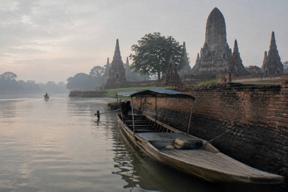 A longtail boat resting on the wide river beside the riverside walls of an ancient temple