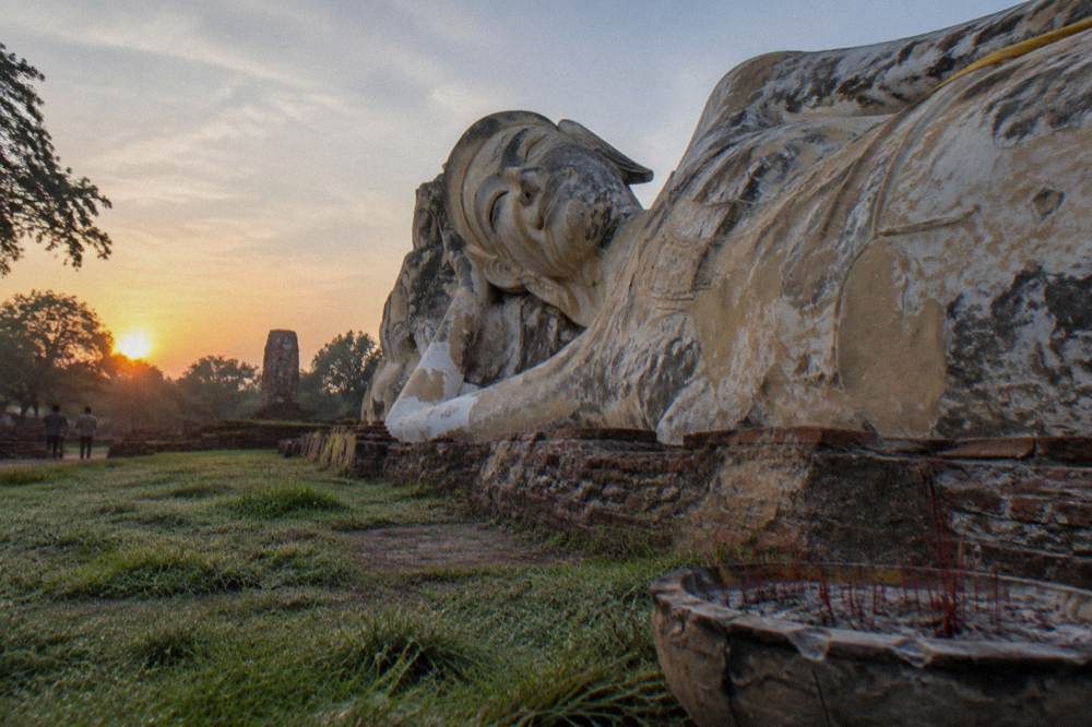 A large stone reclining Buddha resting on open ground beside ruined brick foundations