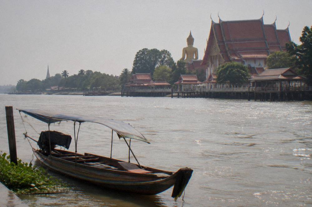 A traditional longtail boat resting on a wide river beside temple ruins on the bank